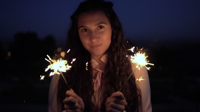 A-young-girl-with-long-dark-hair-stands-against-the-background-of-the-city-at-night-and-holds-fireworks-in-her-hands.-slow-motion.-4K