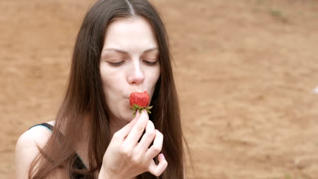 Young-woman-brunette-eats-a-strawberry-sitting-on-the-beach.-Face-close-up.