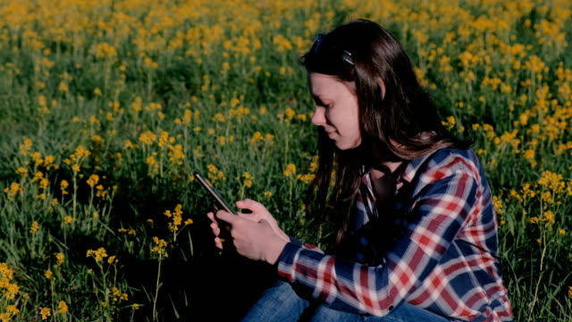 Woman-looking-at-mobile-phone-sitting-in-Park-on-grass-among-yellow-flowers.