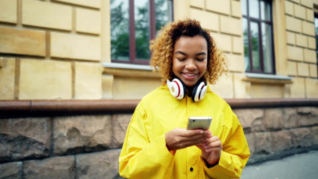 Glücklich-afroamerikanische-Schüler-nutzt-Smartphone-Surfen-oder-SMS-Freunde-stehen-im-freien-in-den-Straßen-der-modernen-Stadt-tragen-von-Kopfhörern-und-lächelnd.