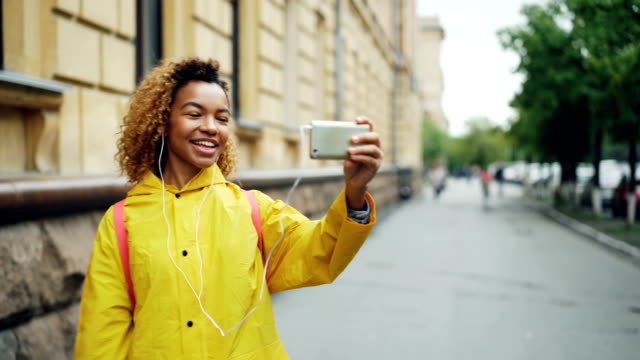 Cute-African-American-teenager-is-talking-to-friends-online-skyping-using-smartphone-and-looking-at-screen-standing-outdoors-in-the-street-and-wearing-earphones.