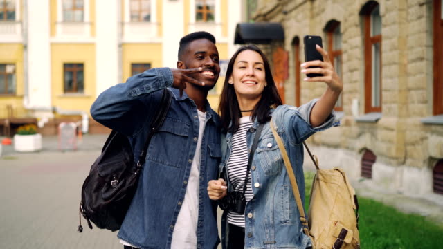 Happy-tourists-friends-with-backpacks-are-taking-selfie-using-smartphone-showing-hand-gestures-v-sign-and-thumbs-up-standing-together-in-the-street-in-beautiful-city.