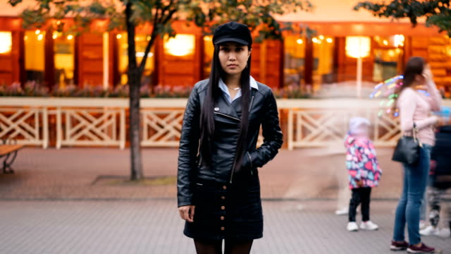 Zoom-out-time-lapse-of-lonely-Asian-lady-looking-at-camera-standing-outside-in-the-street-late-in-the-evening-and-waiting-then-leaving.-People-are-passing-by-quickly.