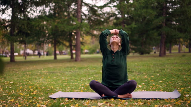 Pretty-young-woman-is-finishing-yoga-practice-sitting-on-mat-closing-eyes-and-breathing-on-peaceful-autumn-day-in-park.-Recreational-activity-and-urban-people-concept.