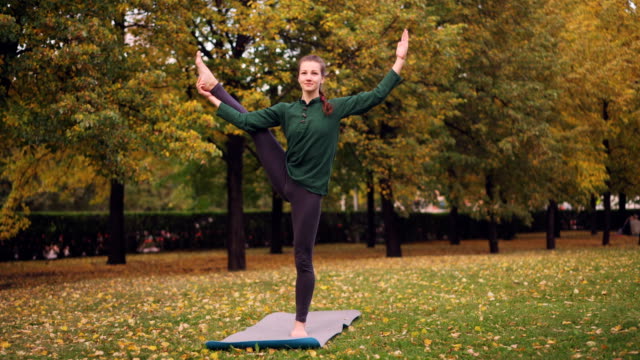 Smiling-girl-professional-yoga-instructor-is-doing-balancing-exercises-standing-on-one-leg-on-mat-on-grass-in-park.-Beautiful-autumn-nature-is-visible.