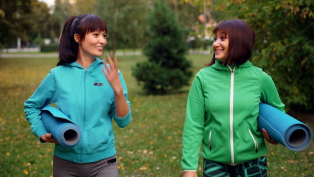 Dolly-Schuss-gut-aussehende-Sportlerinnen-sprechen,-Lächeln-und-Yoga-Matten-zu-Fuß-im-Park-auf-Herbsttag-hält.-Urbane-Menschen,-Freizeit-und-Sport-Konzept.