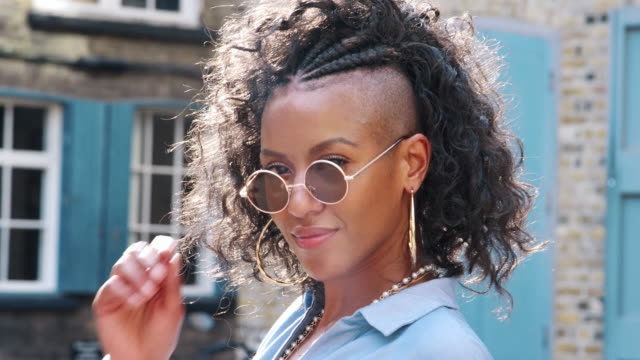 Trendy-young-black-woman-in-blue-dress-and-sunglasses-turns-to-look-to-camera,-close-up,-back-view