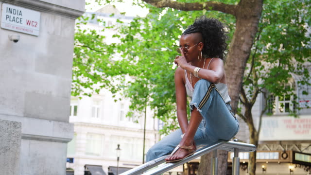 Trendige-junge-schwarze-Frau-tragen-Leibchen-und-Seite-Streifen-Jeans-sitzt-am-Handlauf-in-Stadt-Straße-sprechen-passt-ihre-Sonnenbrille