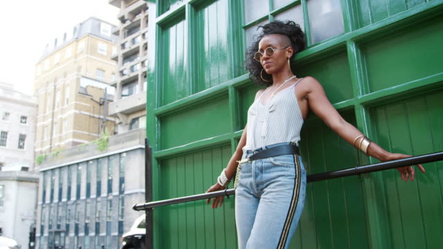Trendy-young-black-woman-waring-round-sunglasses,-camisole-and-jeans-leaning-on-a-handrail-in-a-city-street,-low-angle