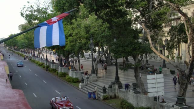 ángulo-alto-establecimiento-de-tiro-de-coche-americano-clásico-en-la-calle-con-la-bandera-cubana-en-la-Habana,-Cuba