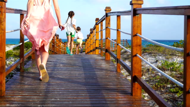 Back-view-family-mother-and-daughters-walking-on-wooden-deck-near-the-beach-enjoying-tropical-summer-vacation.