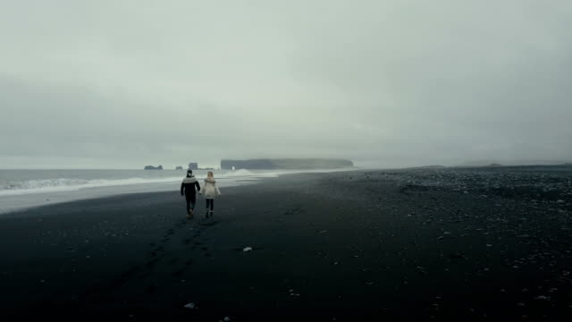 Hintere-Luftaufnahme-des-jungen-Paares-in-isländische-Pullover-zu-Fuß-am-Ufer-des-Meeres-auf-schwarzen-Vulkanstrand-in-Island