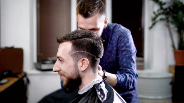 Side-view-of-a-head-of-stylish-smiling-bearded-man-sitting-in-the-barbershop-covered-with-black-peignoir.-Barber-in-casual-clothes-standing-behind-customer-making-boxer-haircut-with-clipper-and-comb