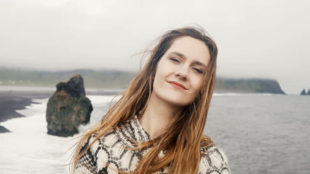 Porträt-der-jungen-glücklich-Frau-stand-am-schwarzen-Strand,-Blick-auf-Kamera-und-lächelnd,-in-der-Nähe-von-Troll-Zehen-Felsen-in-Island