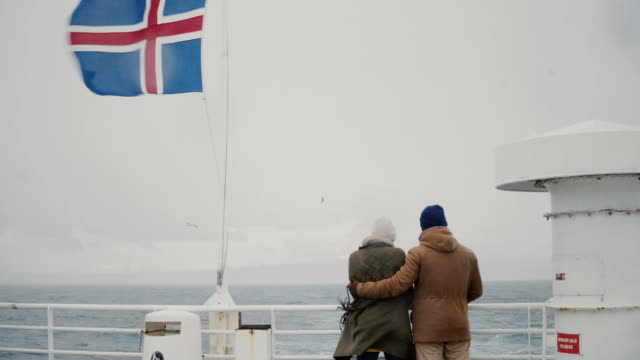 Vista-posterior-de-pie-de-la-joven-pareja-elegante-en-el-tablero-de-la-nave-con-bandera-islandesa.-Hombre-y-la-mujer-miran-el-mar