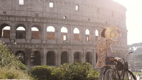 Beautiful-young-woman-in-colorful-fashion-dress-alone-on-hill-with-bike-looking-at-colosseum-in-Rome-at-sunset-with-trees-attractive-girl-with-elegant-straw-hat