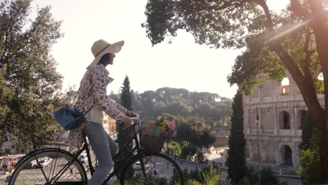Beautiful-young-woman-in-colorful-fashion-riding-bike-in-front-of-colosseum-in-Rome-at-sunset-with-trees-happy-attractive-tourist-girl-with-straw-hat-in-colle-oppio-steadycam-dolly