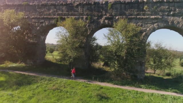 Hombre-joven-mochilero-caminando-por-camino-de-tierra-a-lo-largo-del-antiguo-acueducto-romano-en-naranja-ropa-deportiva-senderismo-dolly-de-drone-de-vista-aérea