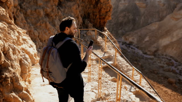 Male-Caucasian-tourist-on-ancient-mountain-road.-Man-with-smartphone-and-camera-enjoys-historic-desert-Israel-ruins.-4K