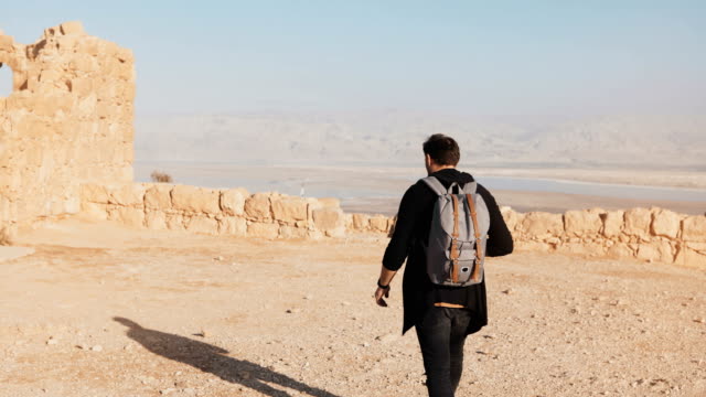 Man-with-backpack-walks-on-desert-mountain-top.-Casual-journalist-blogger-takes-photos-of-Dead-Sea-scenery.-Israel-4K