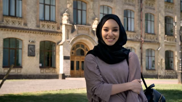 Young-sweet-muslim-girl-in-hijab-is-standing-and-smiling-in-daytime-in-summer,-watching-at-camera,-building-on-background,-religiuos-concept