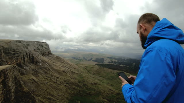 bärtige-Hipster-Mann-nimmt-Bilder-auf-seinem-Smartphone-während-auf-den-Felsen-in-den-Bergen-des-Kaukasus