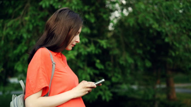 Seitenansicht-der-attraktive-schwangere-Frau-im-Park-spazieren-und-mittels-Smartphone,-beobachtet-Person-Bildschirm-dann-schaut-sich-um-die-schöne-Natur-genießen.