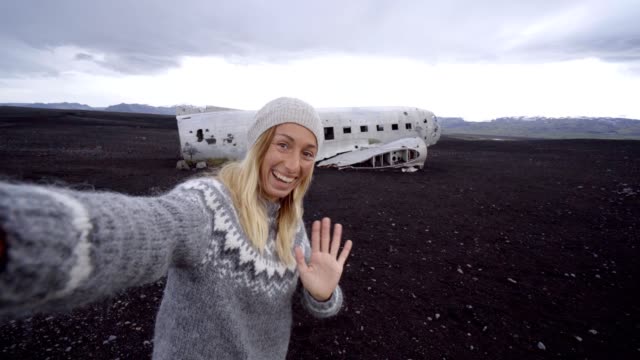 4K-joven-por-ruina-del-aeroplano-en-la-playa-de-arena-negra,-produciendo-una-selfie-retrato-famoso-para-visitar-en-Islandia-y-posan-junto-a-los-restos-del-naufragio