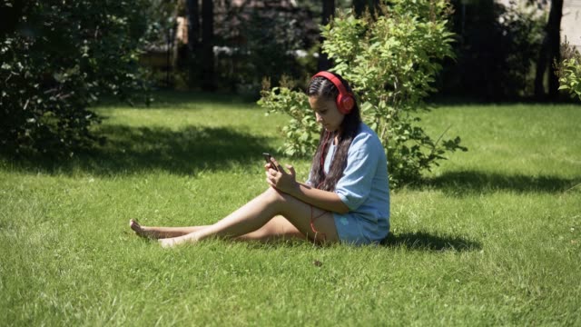 Chica-joven-escuchando-música-con-auriculares-sentado-sobre-la-hierba-en-el-parque.-4K