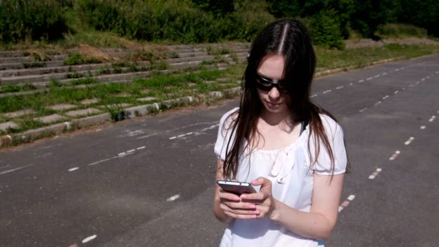 Young-brunette-girl-walking-in-the-stadium-and-typing-a-message-on-a-mobile-phone-and-smiling.