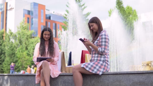 Zwei-attraktive-Mädchen-nach-dem-Einkauf-sitzen-in-einem-Park-nahe-dem-Brunnen-bei-sonnigem-Wetter-mit-ein-Telefon-und-ein-Tablett-mit-einer-guten-Stimmung.-4K