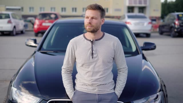 An-attractive-man-with-a-beard-stands-next-to-his-car-in-the-parking-lot.-He-is-thoughtful,-and-looks-away,-someone-is-waiting