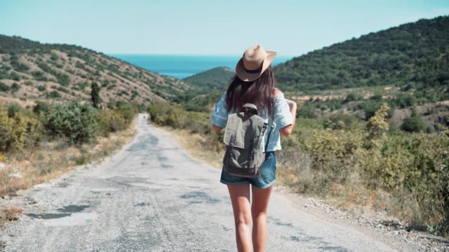 Mujer-de-backpacker-activo-de-vista-posterior-con-sombrero-caminando-en-camino-al-mar-rodeado-de-montañas