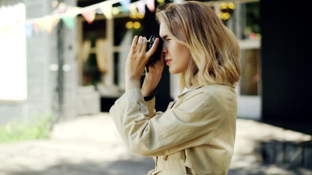 Cheerful-young-girl-is-adjusting-camera-then-taking-photos-of-beautiful-city-standing-outdoors-in-the-street.-Hobby,-occupation,-modern-technology-concept.