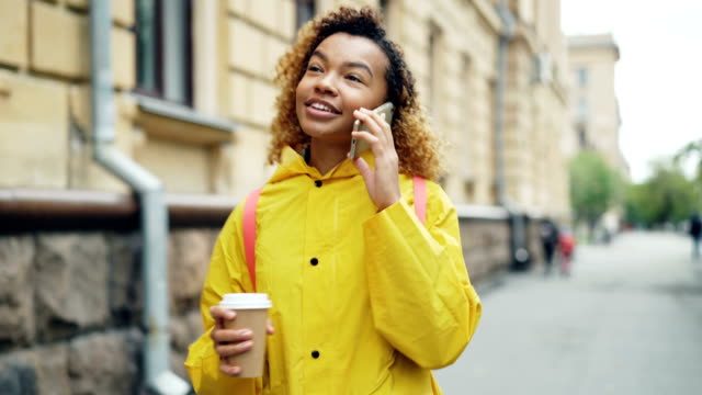 Cheerful-mixed-race-teenage-girl-is-talking-on-smart-phone-and-holding-take-out-coffee-walking-along-street-in-beautiful-city.-Modern-lifestyle-and-technology-concept.