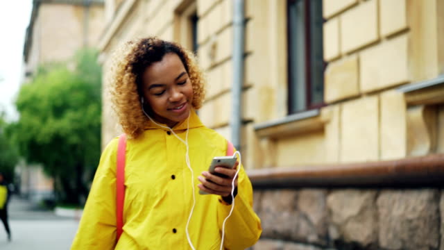 Good-looking-African-American-lady-is-listening-to-the-radio-wearing-earphones-and-holding-modern-smartphone,-girl-is-singing-and-enjoying-music-and-rhythm.