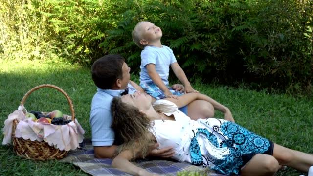 Family-picnic-in-nature.-Mom,-dad-and-son-lie-on-the-grass-in-the-Park-near-the-fruit-basket.-They-look-up,-laugh,-play-and-have-fun.-Boy-eating-grapes.