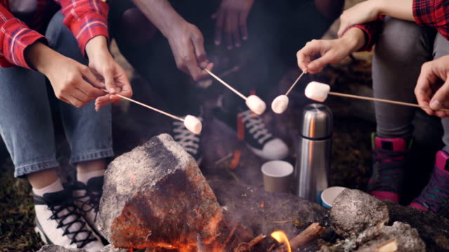 Close-up-shot-of-burning-campfire-and-people's-hands-holding-sticks-with-marshmallow-above-flame-and-tourists'-legs-getting-warm-near-fire.-Camping-and-food-concept.