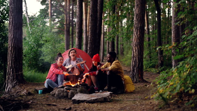 Man-Touristen-in-Freizeitkleidung-spielt-Gitarre,-während-seine-Freunde-kochen-und-Essen-süßen-Marshmallow-auf-Herbsttag-Lagerfeuer-sitzen.-Menschen-und-Camp-Konzept.