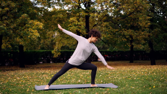 Beautiful-girl-with-short-curly-hair-is-doing-yoga-outdoors-standing-in-Warrior-pose-then-stretching-body-and-legs-during-practice-in-park.-Trees-and-grass-are-visible.
