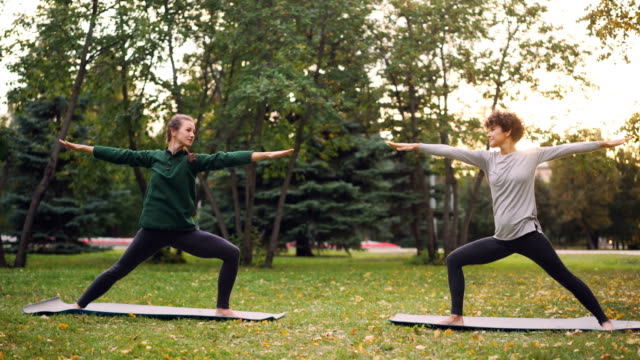 Attractive-girls-are-enjoying-outdoor-yoga-in-park-practising-positions-standing-on-mats.-Instructor-is-speaking-teaching-student,-both-women-are-relaxed-and-happy.