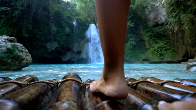 Low-angle-view-of-female's-barefoot-on-bamboo-raft-walking-toward-a-beautiful-waterfall-located-on-Cebu-Island-in-the-Visayas-Islands-in-the-Philippines.-People-travel-sharing-environment-conservation-concept.
