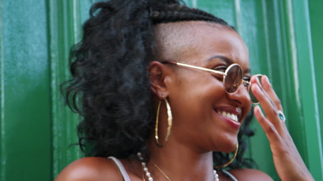 Young-black-woman-wearing-sunglasses-leaning-on-green-wall-in-the-laughing,-head-and-shoulders