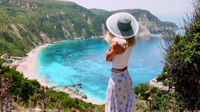 Cute-woman-in-hat-admiring-landscape-on-greek-island.-Blonde-posing-in-front-of-amazing-scenery-of-Petani-bay.-Summer-vacation-lifestyle-travel-adventure-carefree-joy-and-happiness-concept.-4k-video