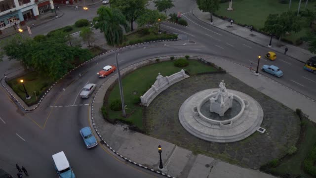high-angle-establishing-shot-of-classic-cars-turning-on-street-roundabout-in-Havana,-Cuba-in-evening