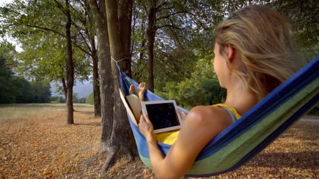 Junge-Frau-in-Hängematte-Sonnenuntergang-Lesung-auf-digital-Tablette-mit-mobilen-Anwendungen-auf-wireless-Technologie.-Die-Leute-reisen-modern-Life-Konzept
