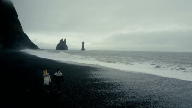 Vista-aérea-de-la-joven-pareja-caminando-en-la-playa-volcánica-negra-cerca-el-troll-del-pie-acantilados-en-Islandia
