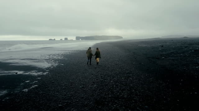 Back-aerial-view-of-young-hipster-couple-walking-on-the-black-volcanic-beach-in-Iceland.-Man-and-woman-enjoy-the-sea