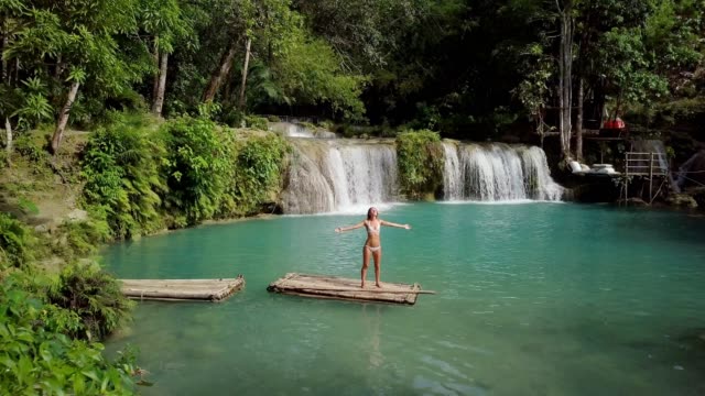 Drone-shot--Young-woman-on-bamboo-raft-embracing-nature-at-waterfall-in-rainforest,-arms-wide-open-People-travel-vacations-concept.-4K-resolution,-Philippines