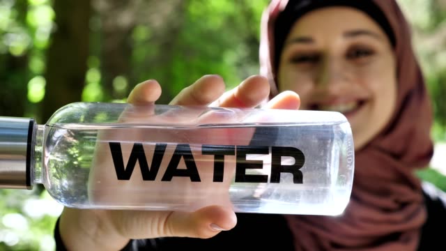 Portrait-of-a-cute-young-girl-in-a-hijab-with-a-bottle-of-water-in-her-hands,-smiling,-looking-at-the-camera,-park-in-the-background,-focus-pull-50-fps